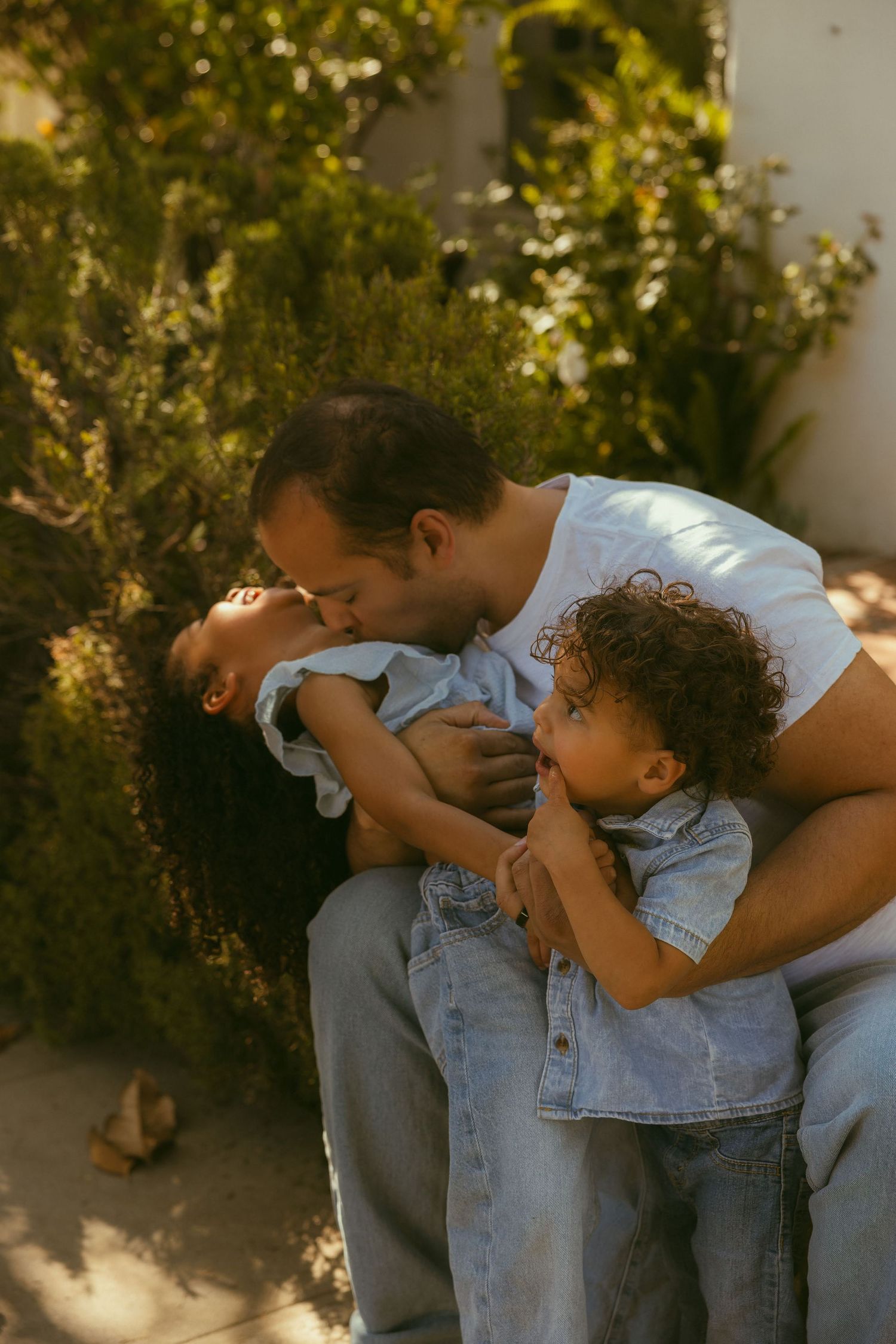 A father kisses his laughing daughter while their curly-haired toddler watches outdoors in warm sunlight.