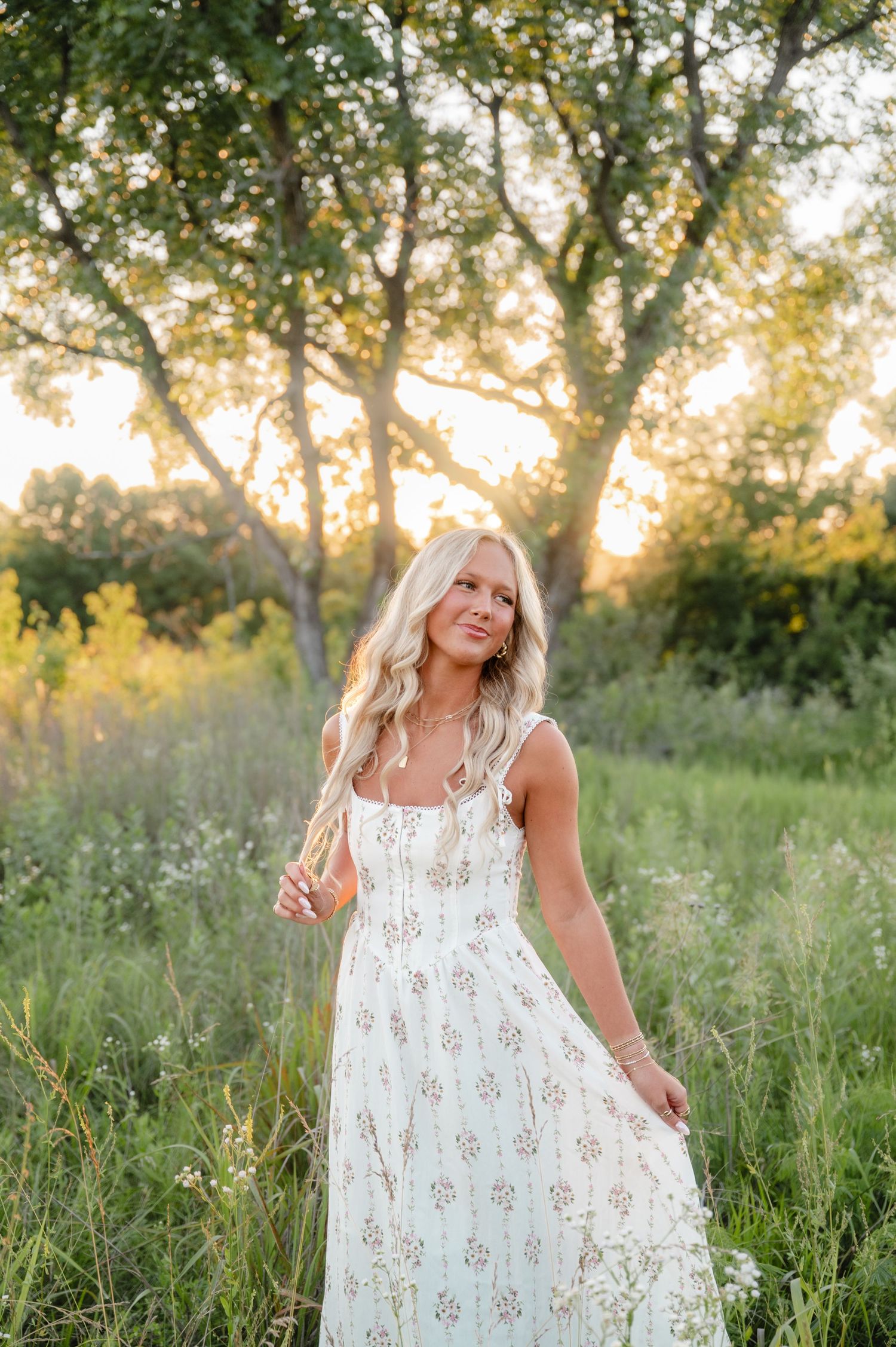 Young blonde woman in a floral white sundress poses in a sunlit meadow with trees glowing in golden hour light.