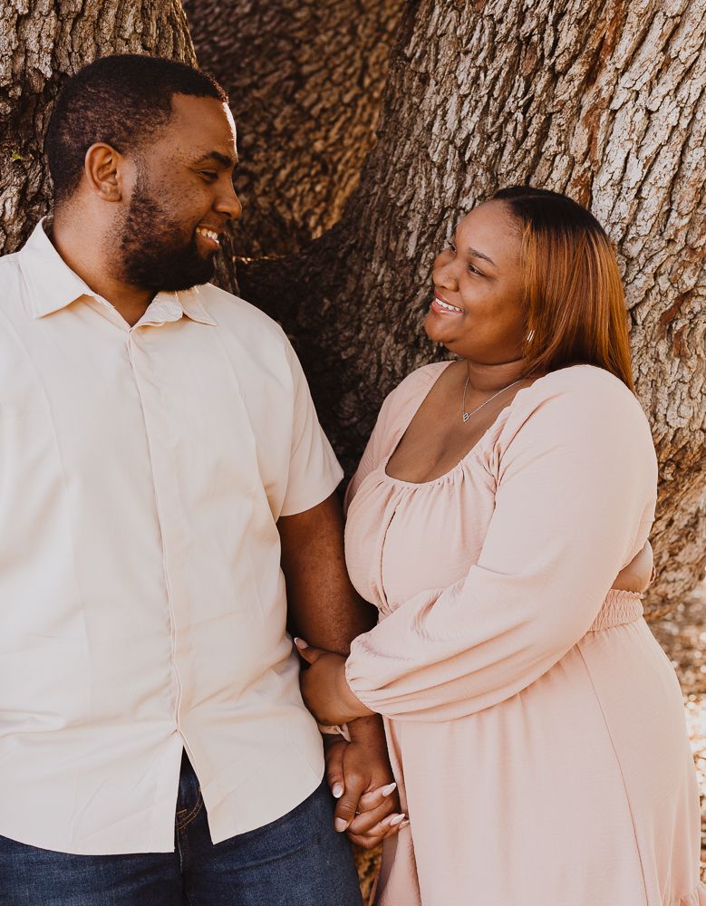 A couple holds hands and smiles at each other while leaning against a large tree outdoors.