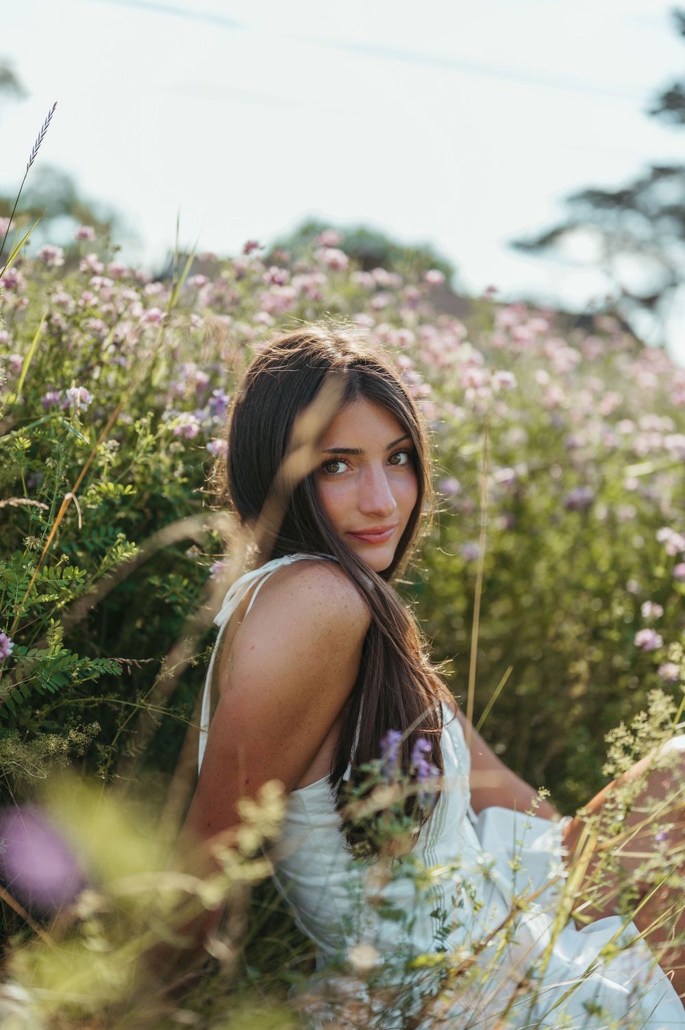 Young woman in white dress sitting among wildflowers and pink blooms, looking over shoulder in golden hour light.