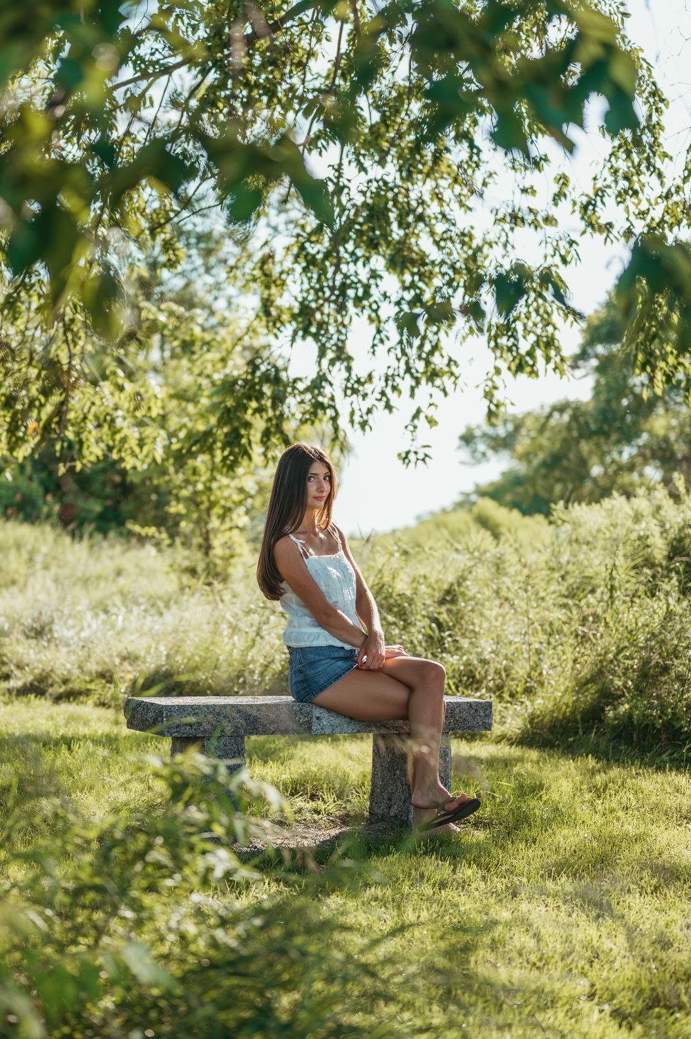 Young woman in denim shorts sitting on a stone bench surrounded by lush green trees in a sunny park.