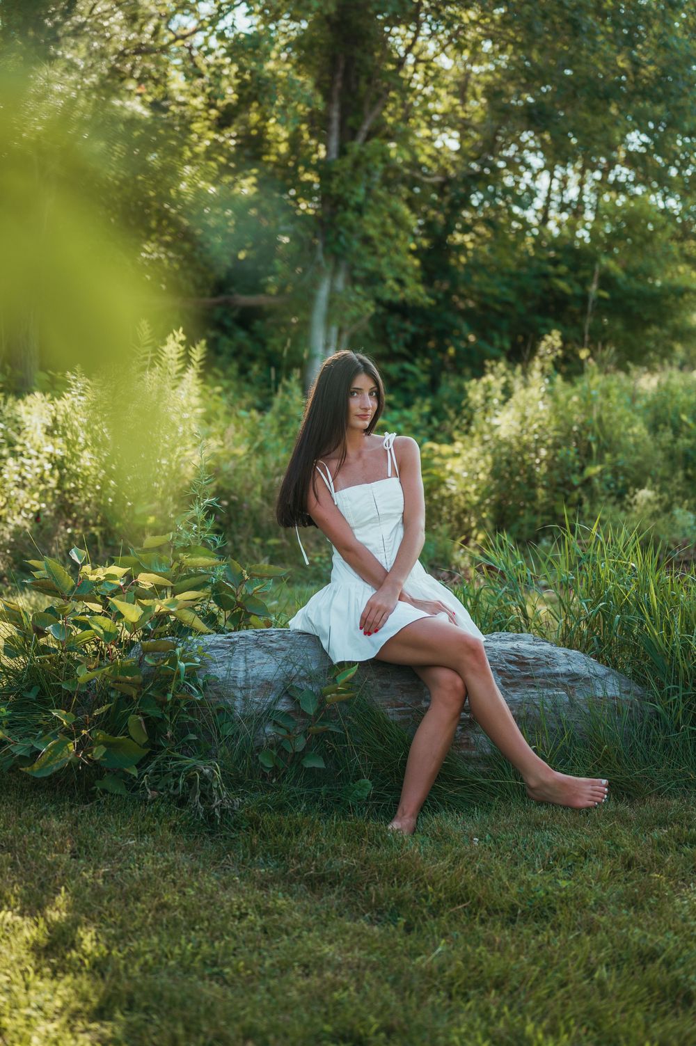 Young woman in white sundress sitting barefoot on a rock surrounded by lush green garden foliage.