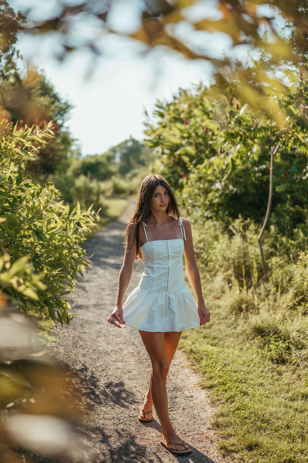 Young woman in white mini dress walking on a sunny dirt path surrounded by lush green trees.