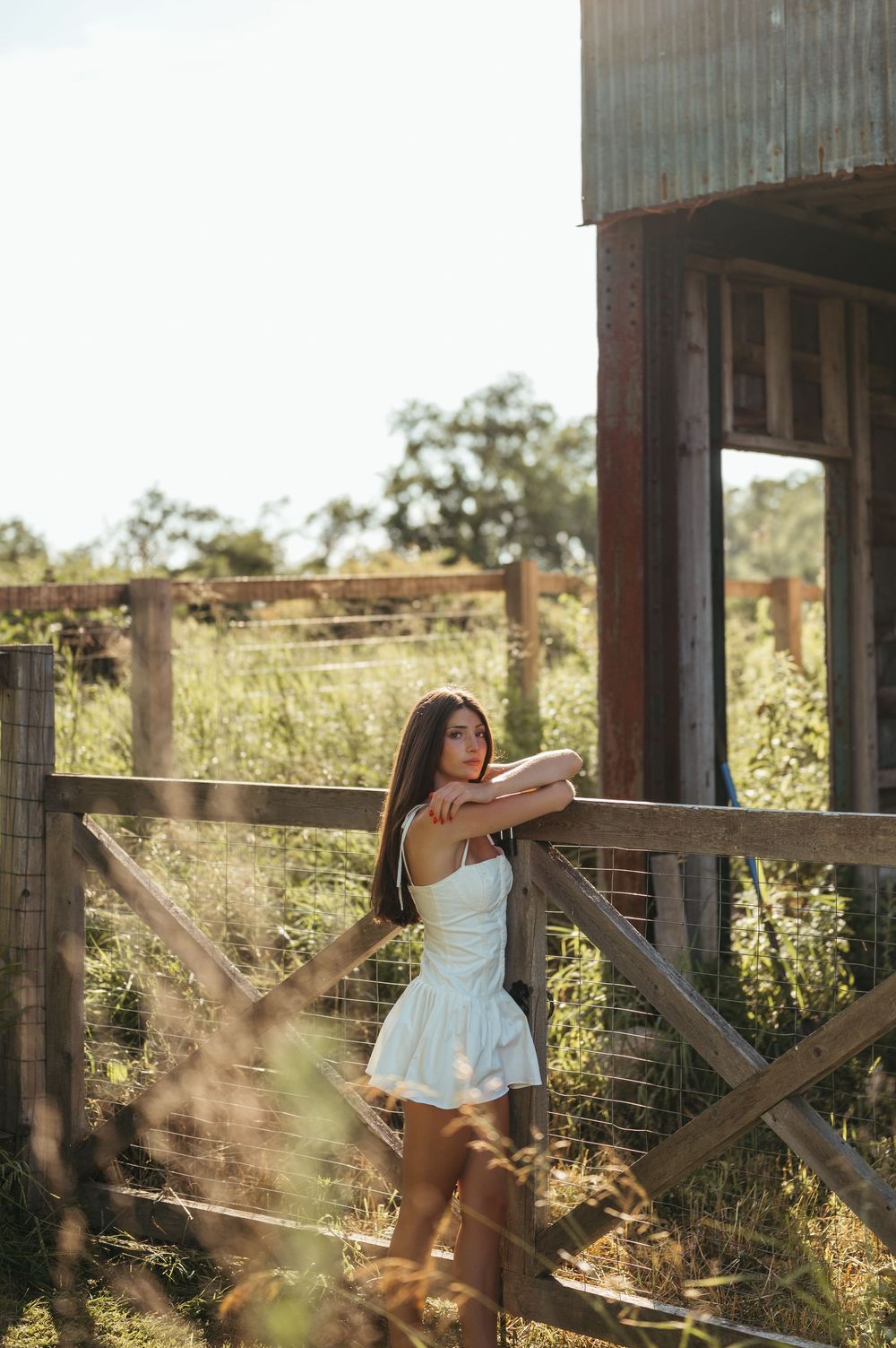 Young woman in white dress leaning on rustic wooden fence near old barn in sunny rural countryside setting.