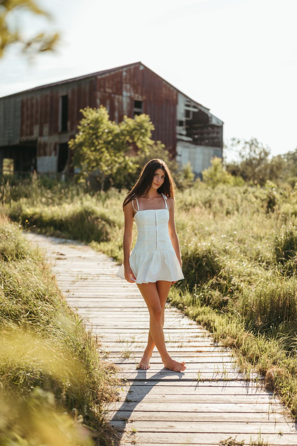 Young woman in white sundress walks barefoot on wooden boardwalk path through green meadow near rustic red barn.