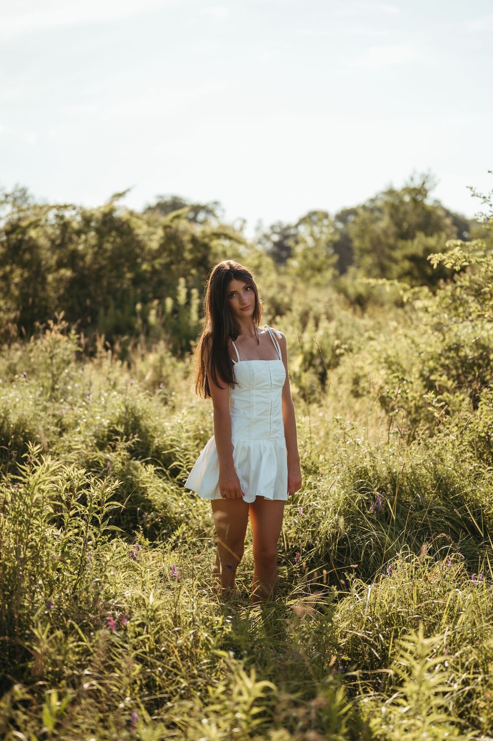 Young woman in white sundress standing in lush green meadow with trees in golden sunlight.