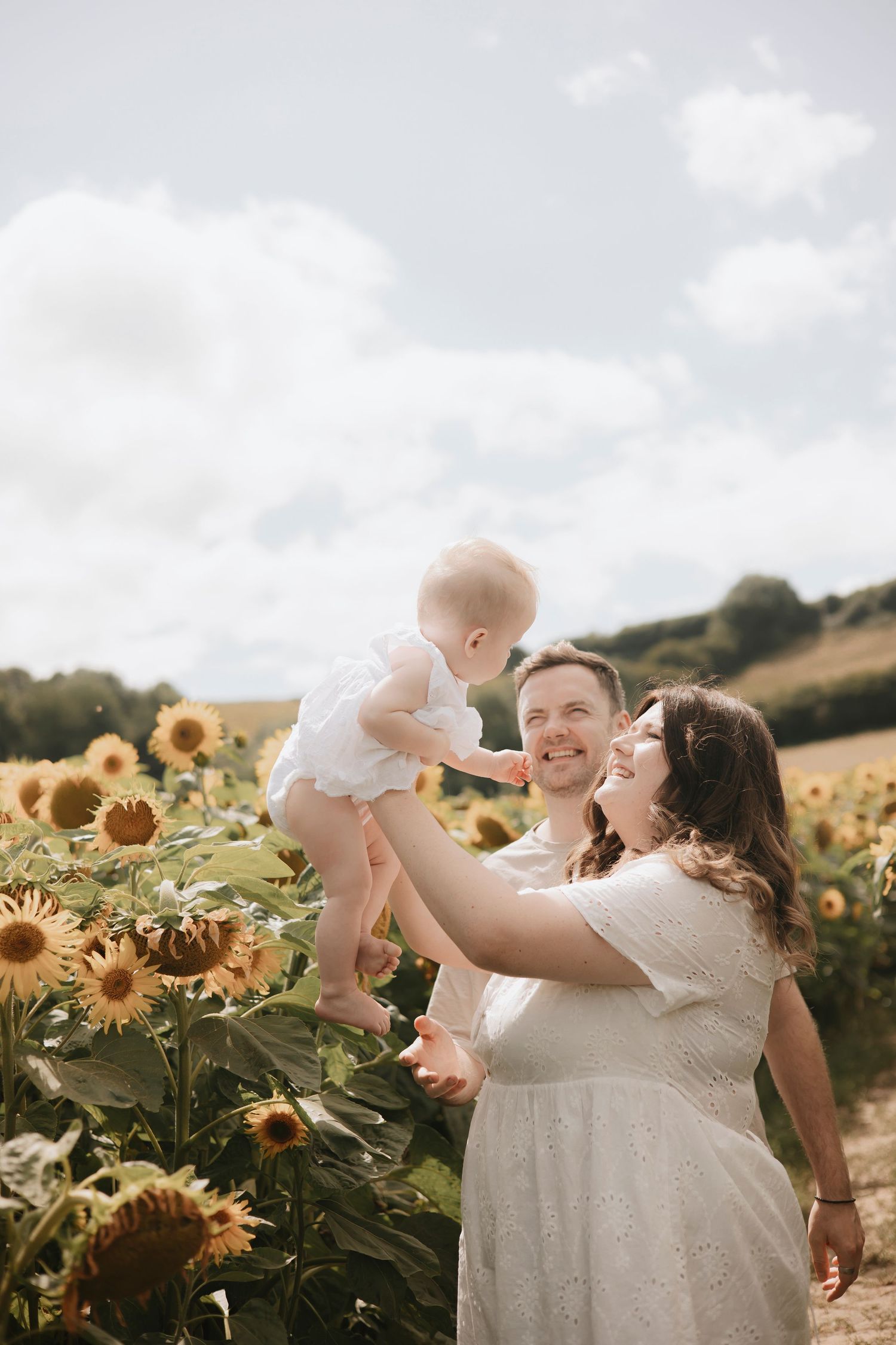Happy family with baby in sunflower field on sunny day, parents smiling and lifting child outdoors.
