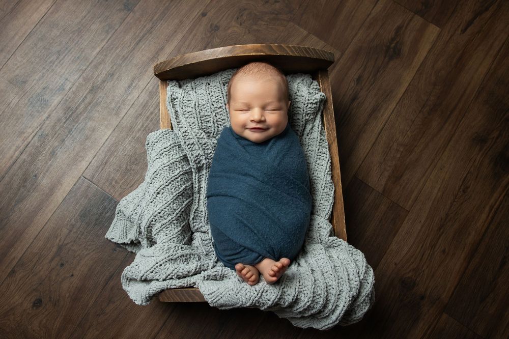 Smiling newborn baby swaddled in teal wrap, resting on a rustic wooden bed with gray knit blanket.