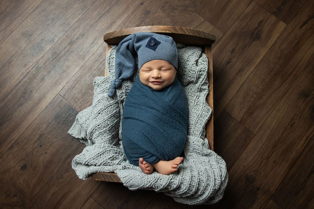 Smiling newborn baby swaddled in blue wrap and hat, sleeping peacefully in a small wooden bed.
