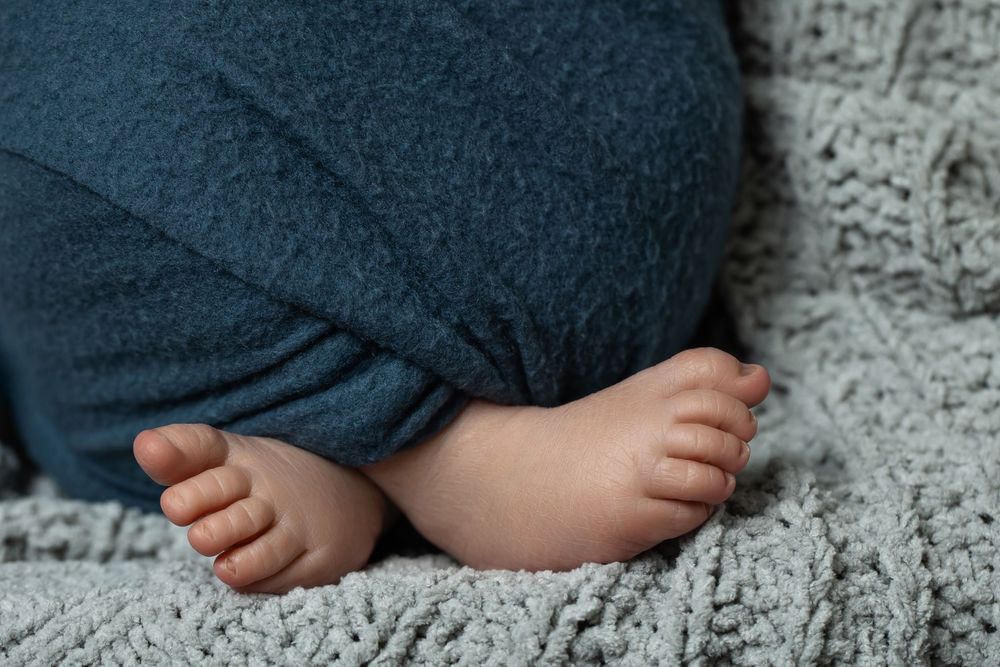 Close-up of tiny newborn baby feet peeking out from a teal blanket on a soft gray knit surface.