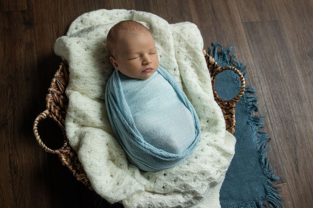 Newborn baby swaddled in blue wrap sleeping peacefully in a wicker basket with a cream knit blanket.