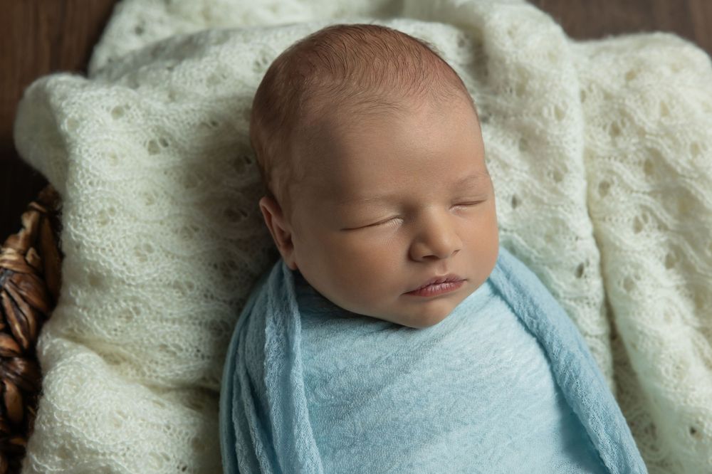 Peaceful newborn baby sleeping wrapped in a soft blue swaddle blanket inside a cozy wicker basket.