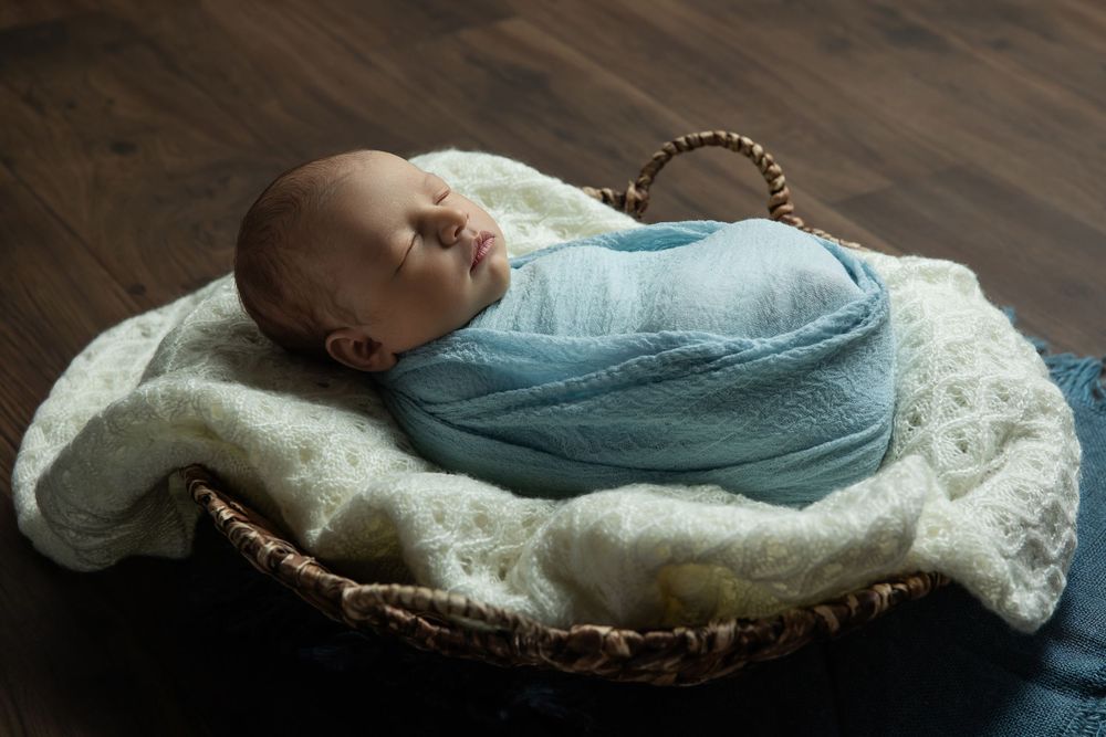 Newborn baby swaddled in blue wrap sleeping peacefully in a wicker basket lined with white fur.