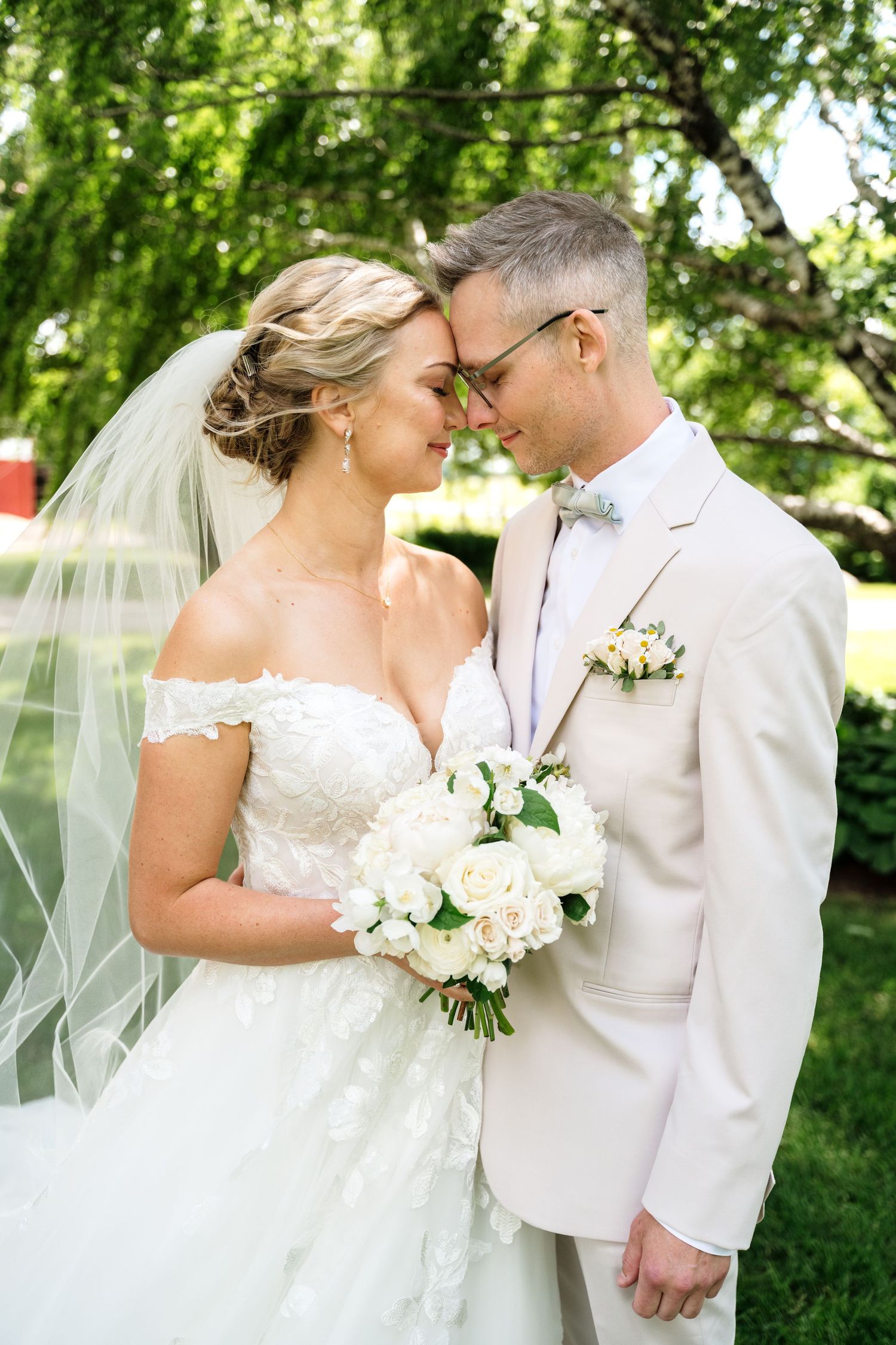 Bride and groom touching foreheads outdoors, bride in lace off-shoulder gown with veil, groom in cream suit with boutonniere.