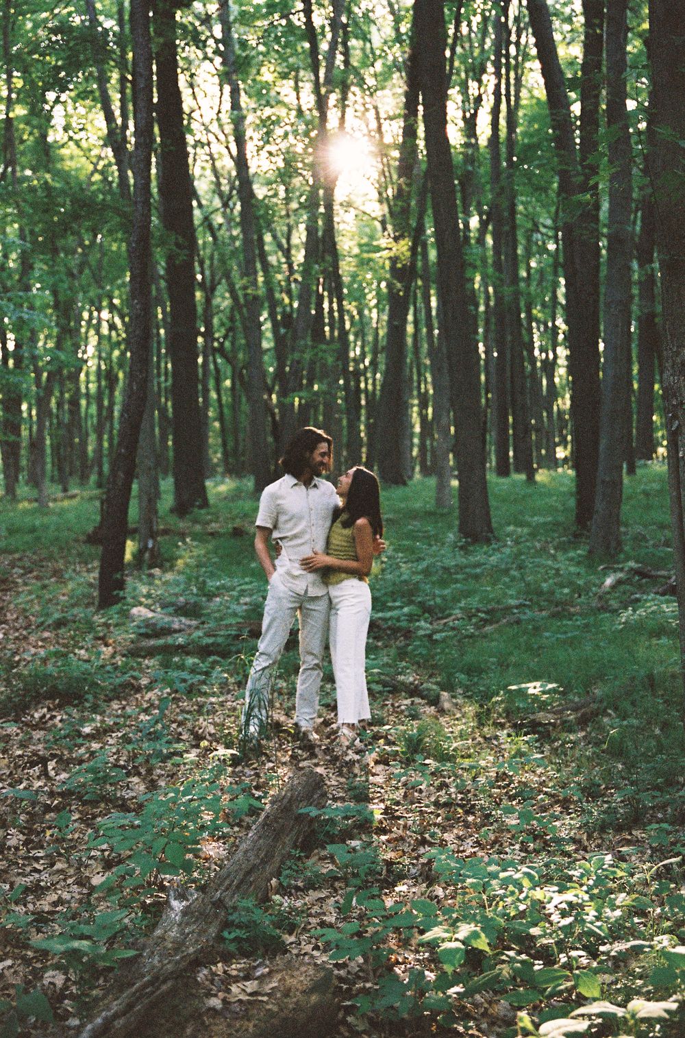 Couple embracing romantically in a sunlit green forest, standing on a log surrounded by lush foliage.
