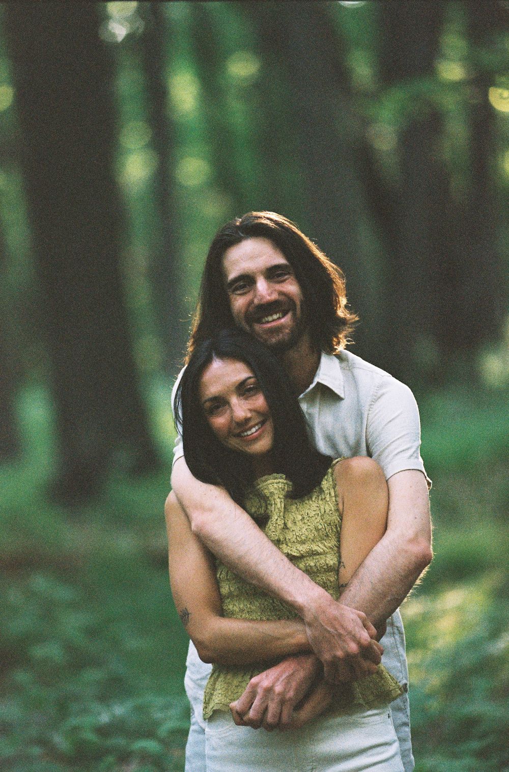 A smiling couple embraces in a lush green forest, the man hugging the woman from behind in a warm, romantic pose.