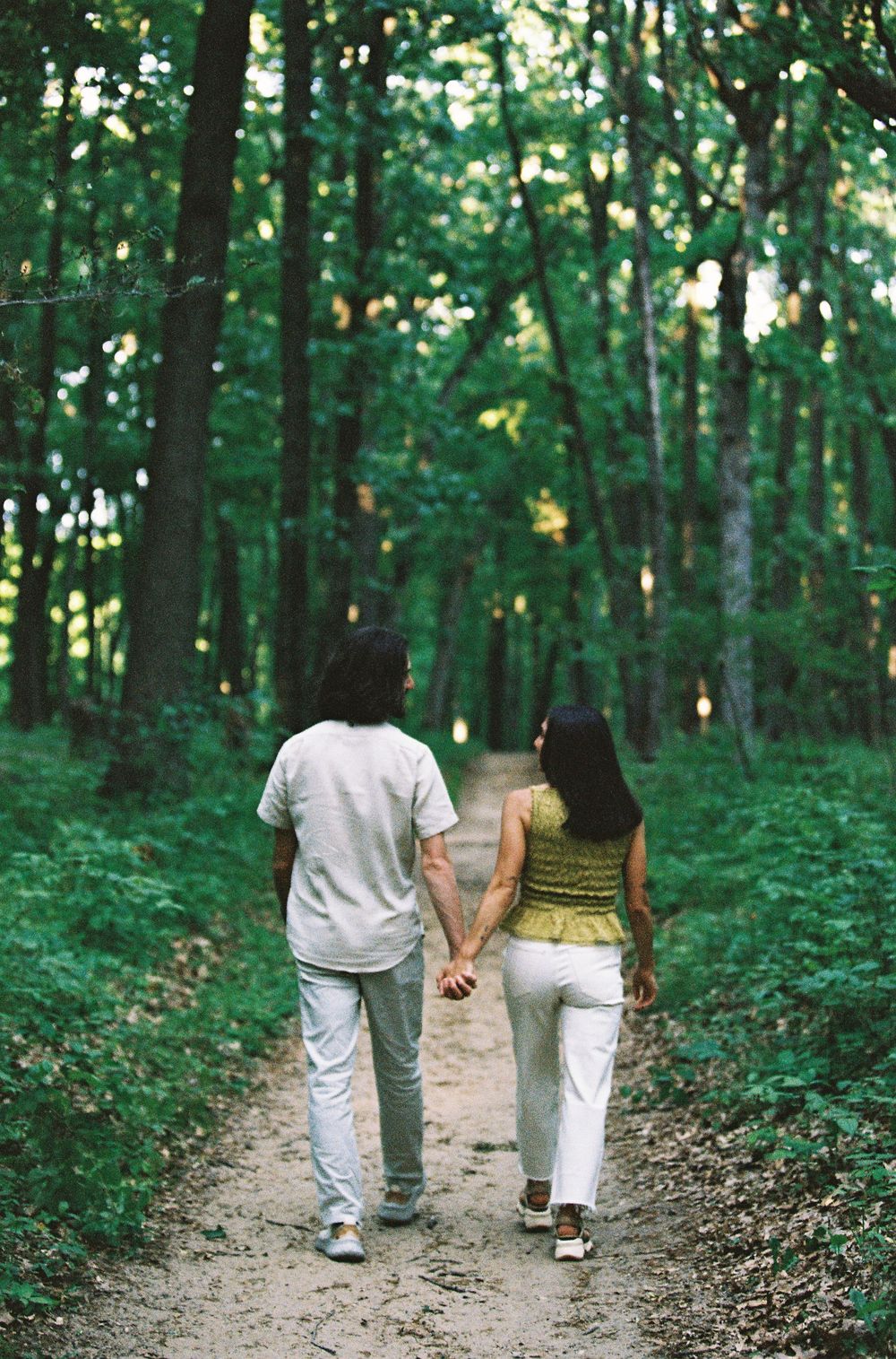 A couple holds hands walking down a forest path surrounded by lush green trees.