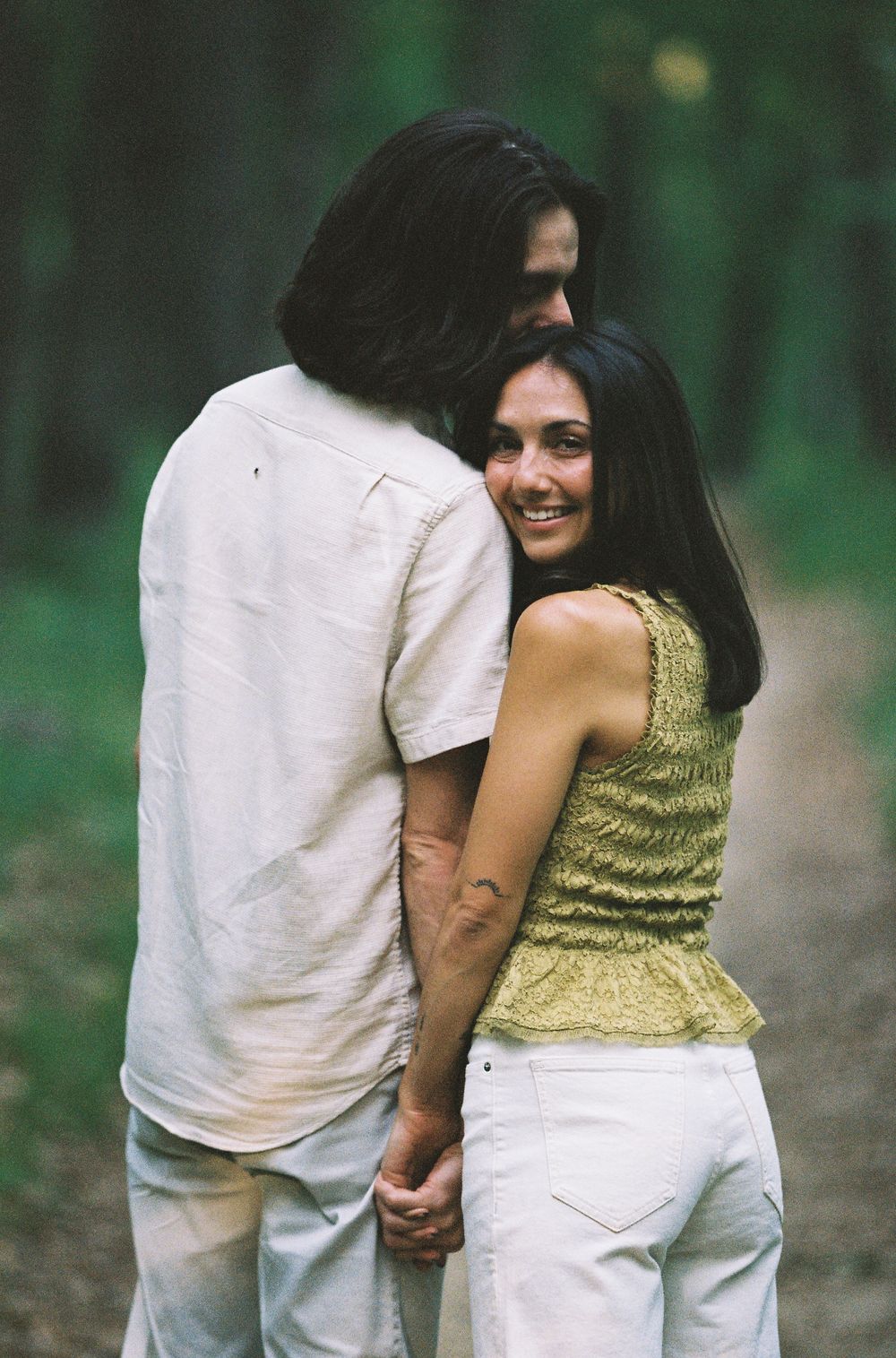 A smiling woman hugs a man from behind on a forest path, both wearing white bottoms in a romantic outdoor setting.