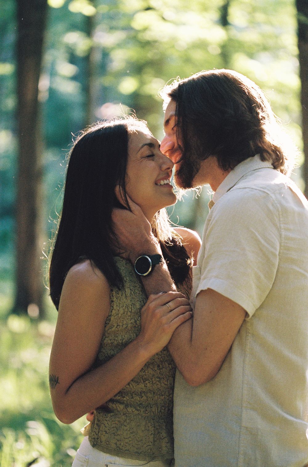 A smiling couple embraces romantically in a sunlit forest, the woman in a green top and man in a white shirt.