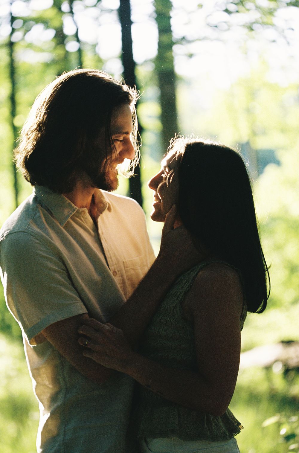 A happy couple embraces and smiles at each other in a sunlit forest, sharing a romantic moment outdoors.