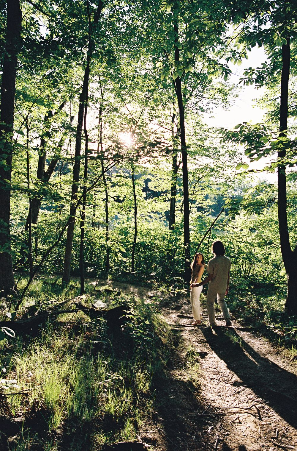 Two people standing on a sunlit forest trail surrounded by lush green trees and foliage.