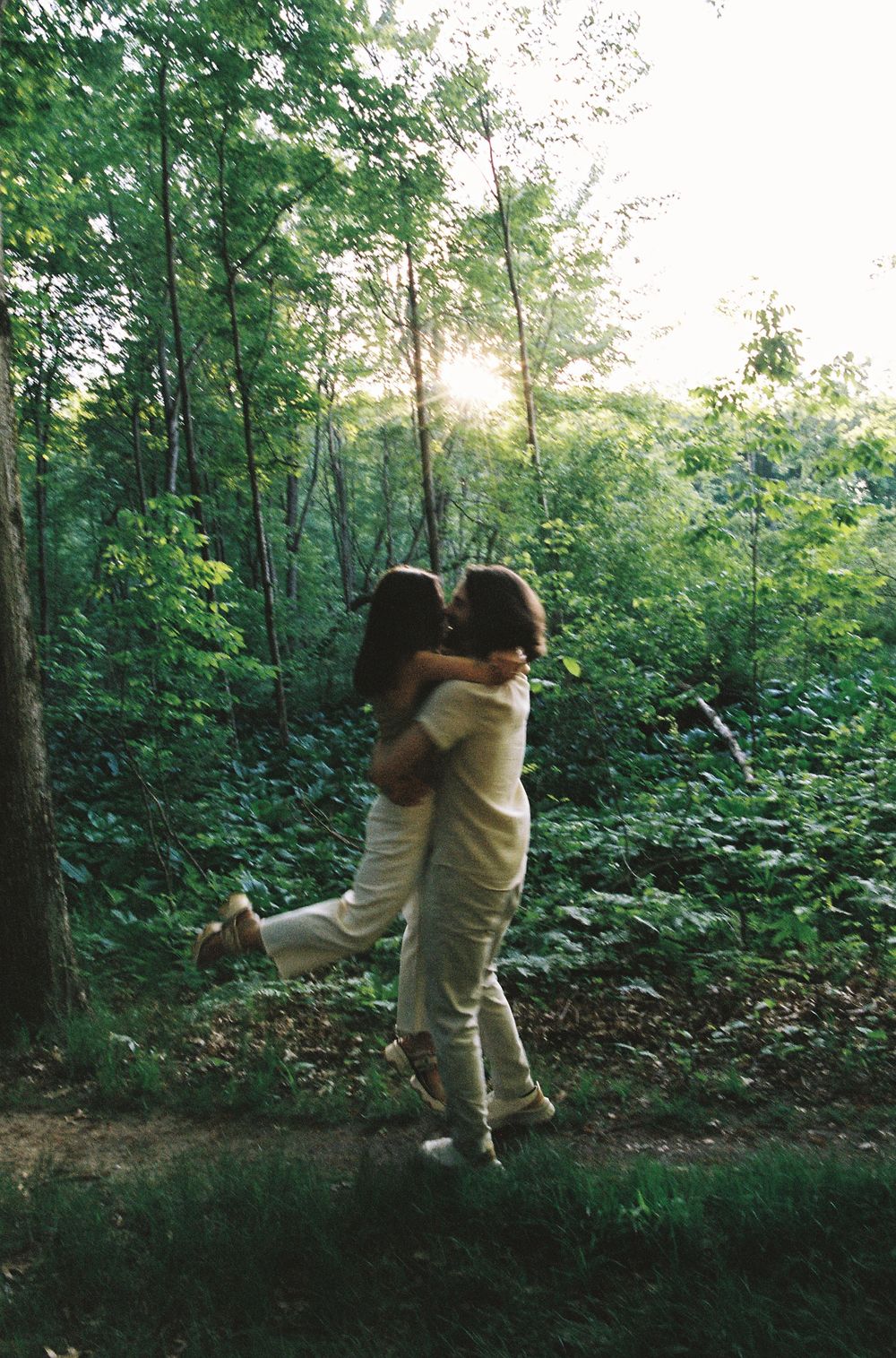 Couple sharing a romantic kiss embrace in a sunlit green forest, surrounded by lush trees and foliage.