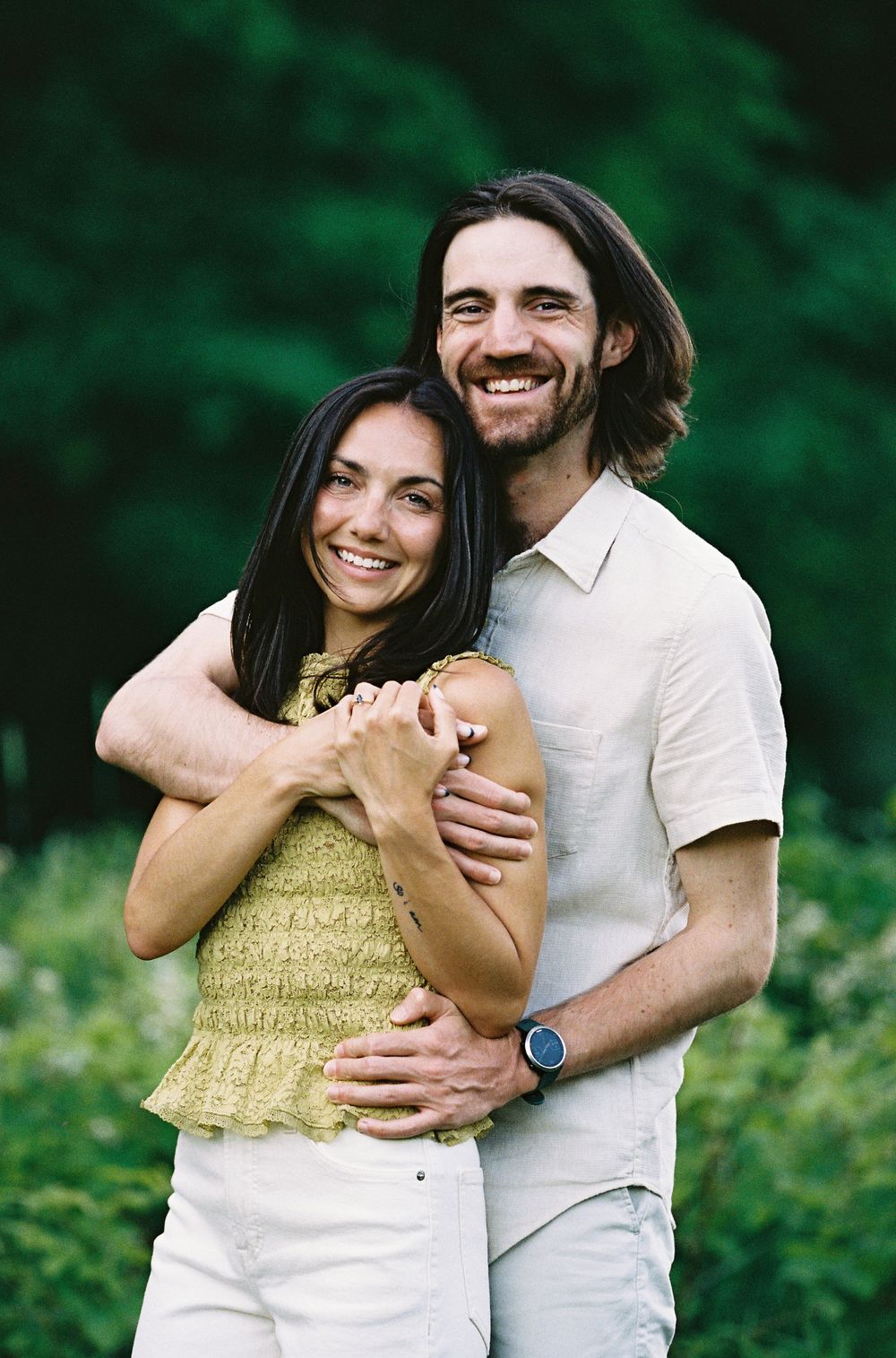 Smiling couple embracing outdoors with lush green trees, woman in yellow top and man in cream shirt wearing a watch.