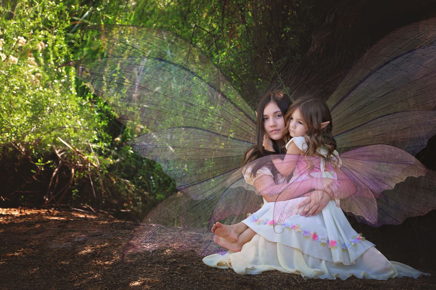A woman and a young girl in white and pink fairy costumes with sheer iridescent wings sit together on a sunlit forest floor.
