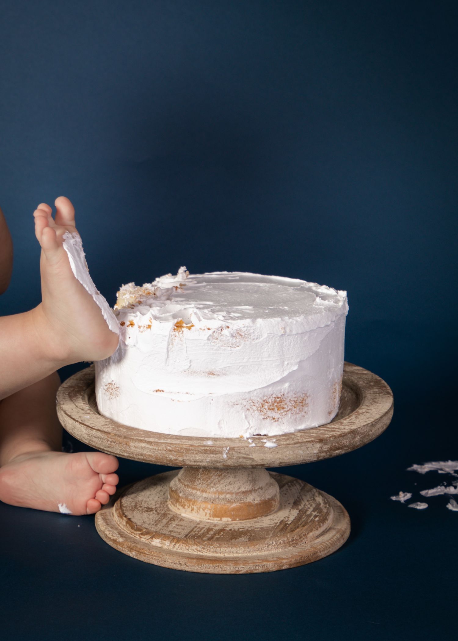A messy baby in denim overalls eating cake with frosting on face and hands against olive background.