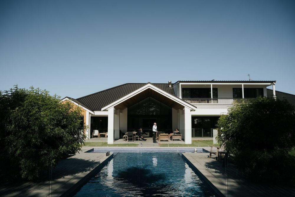 Modern white house with dark roof featuring an outdoor pool, open patio area, and lush green hedges under clear blue sky.