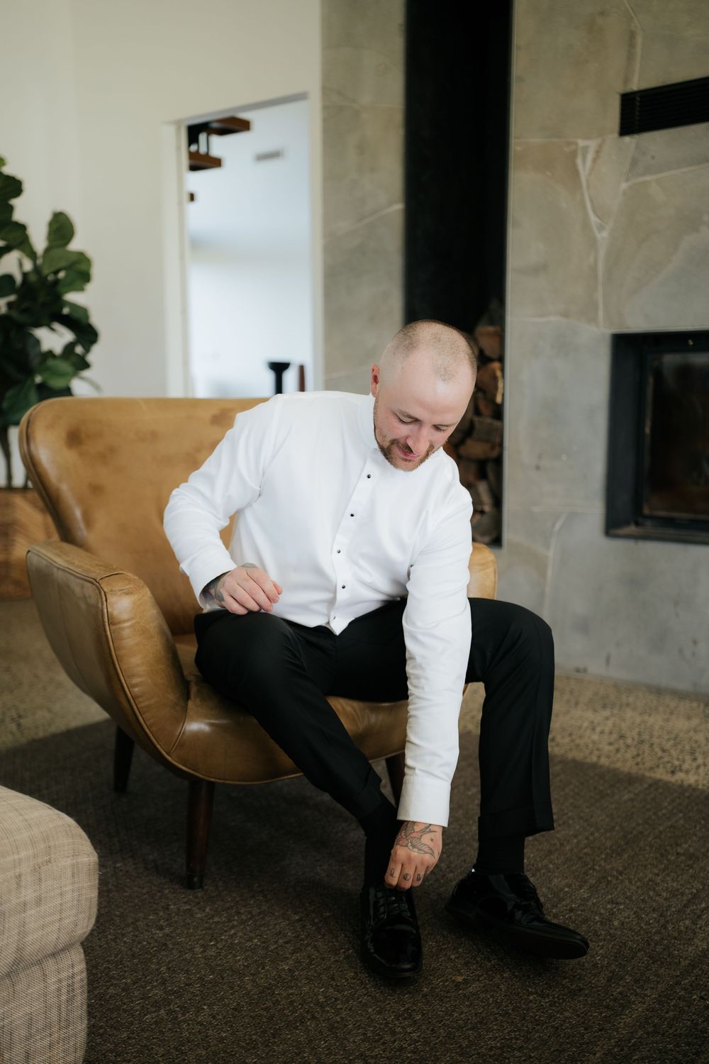 A man in a white dress shirt and black trousers sits in a leather chair, tying his dress shoes near a fireplace.