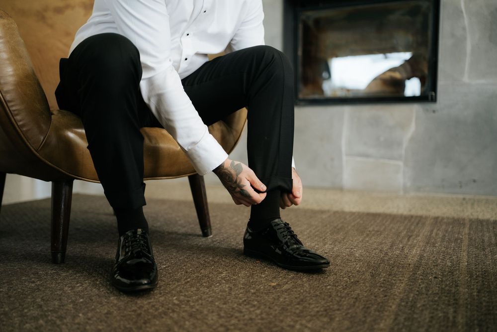 Man in white dress shirt and black pants tying black leather dress shoes while seated in a brown leather chair.