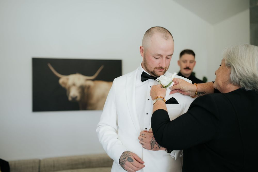 Groom in white tuxedo getting boutonniere pinned by older woman, with bull artwork on wall behind them.