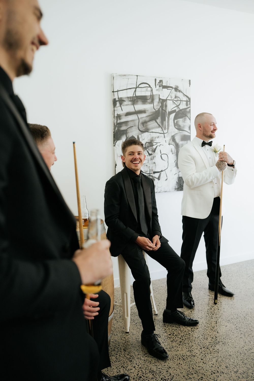 Men in formal black tuxedos laughing and holding billiard cues in a modern art gallery setting.