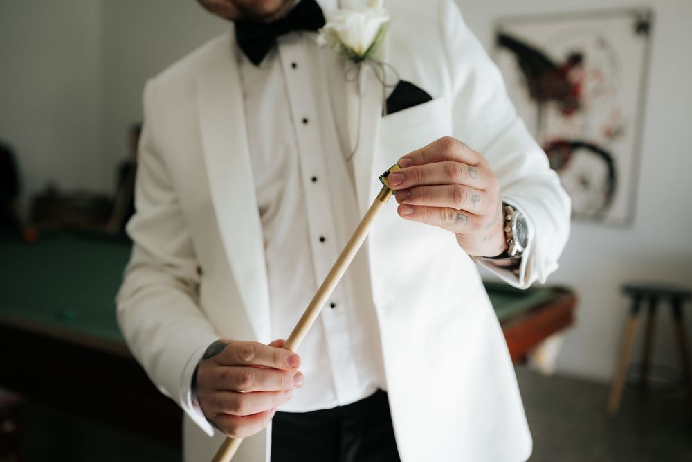 Groom in white tuxedo jacket with bow tie and boutonniere holds a wooden cane, preparing for wedding day.