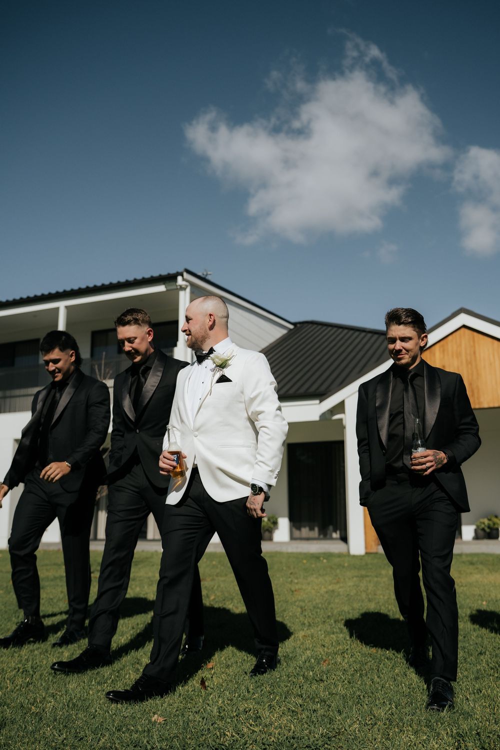 Groom in white tuxedo jacket walks with three groomsmen in black suits on green lawn outside modern venue.