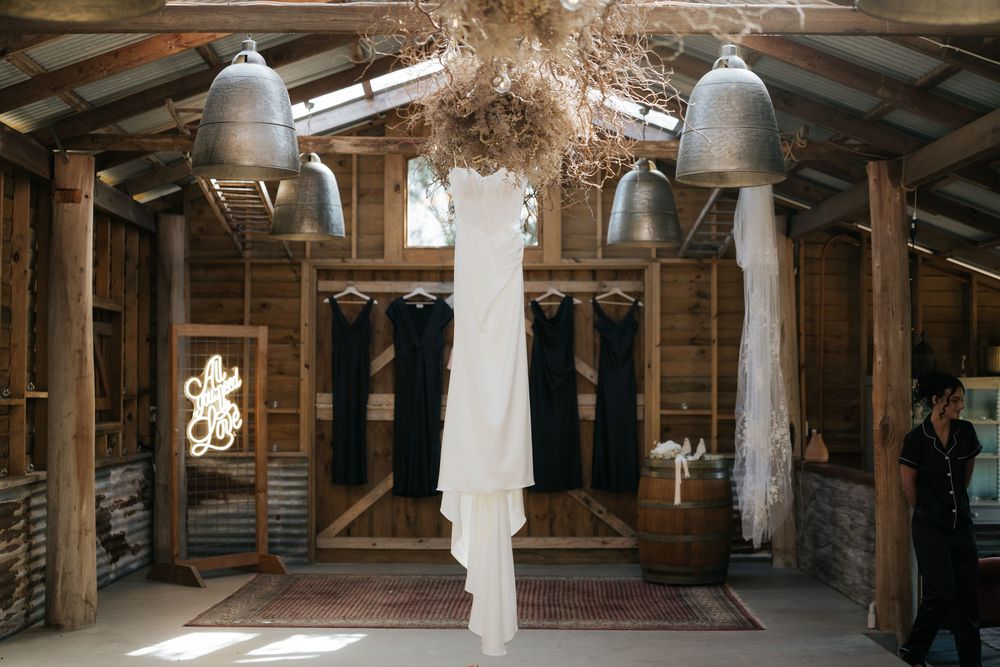 Rustic barn wedding setup with white gown hanging amid dried florals, pendant lights, and dark bridesmaid dresses.