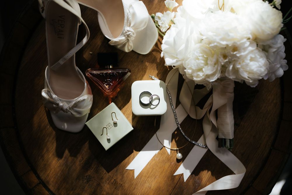 Elegant wedding flat lay featuring bridal heels, white peony bouquet, rings, earrings, and ribbons on wooden surface.