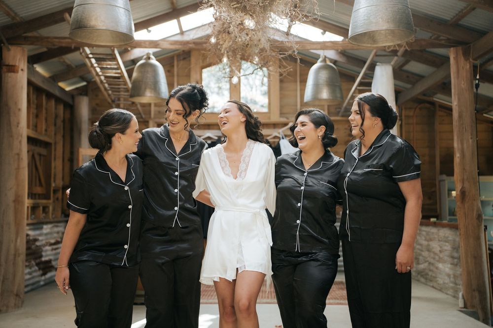 Bride in white robe laughing with bridesmaids in black satin pajamas inside a rustic barn venue.