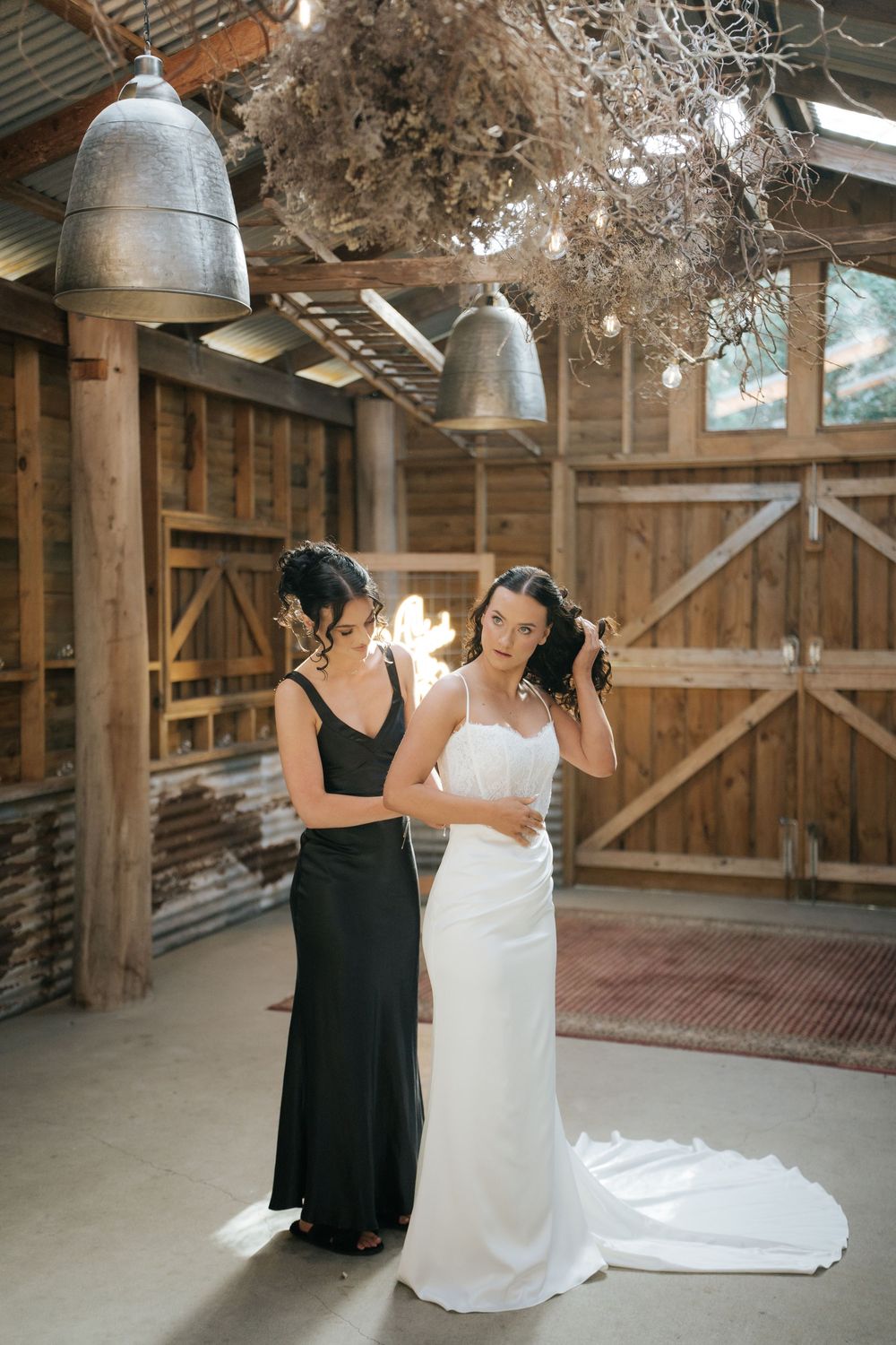 Bride in white gown gets help from bridesmaid in black dress inside rustic barn wedding venue.