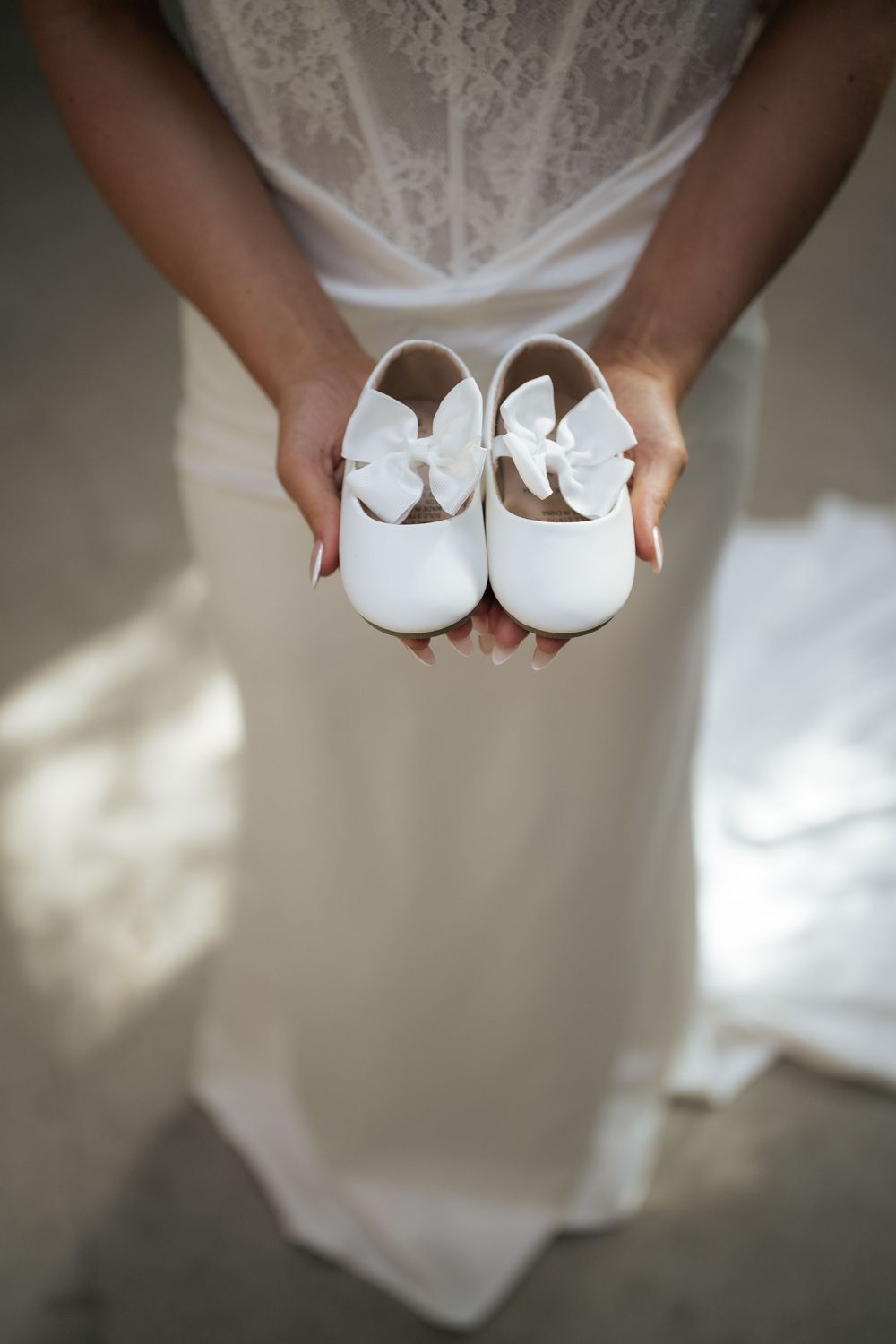 A bride in a white lace dress holds a pair of tiny white baby shoes with bows, symbolizing pregnancy announcement.