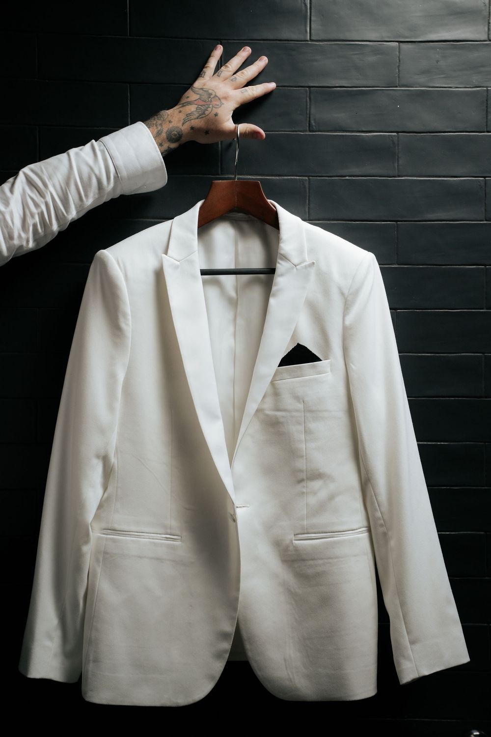 White tuxedo blazer on wooden hanger held by tattooed hand against dark brick wall.