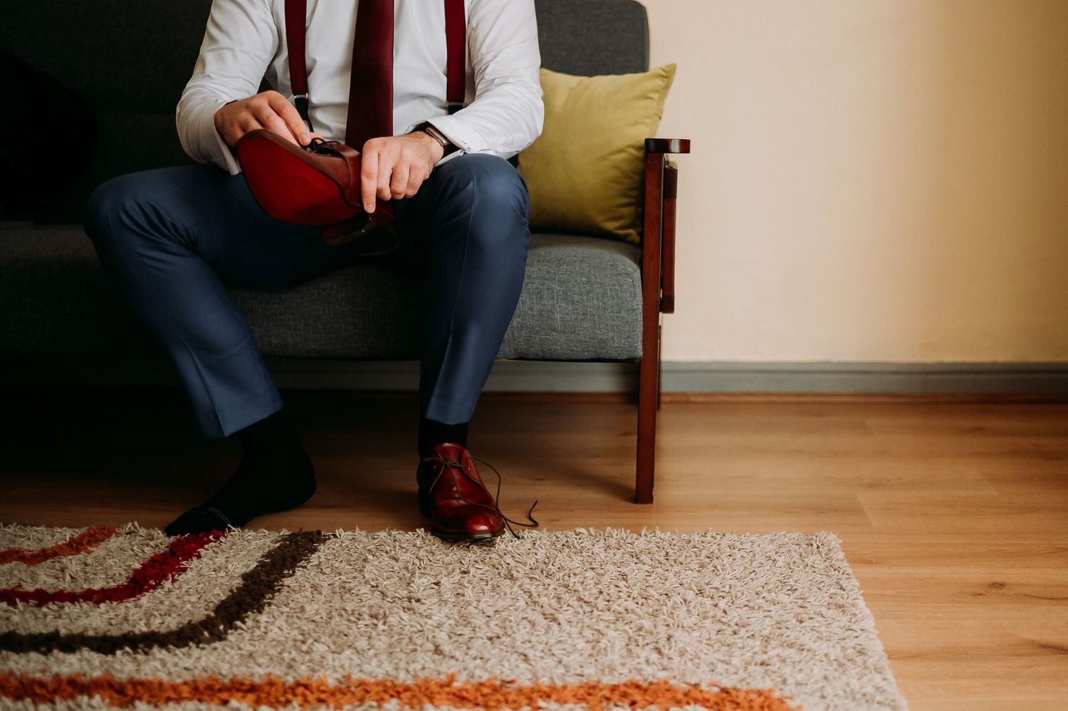 Groom sitting on a sofa holding a wallet, wearing a red tie and brown leather dress shoes.