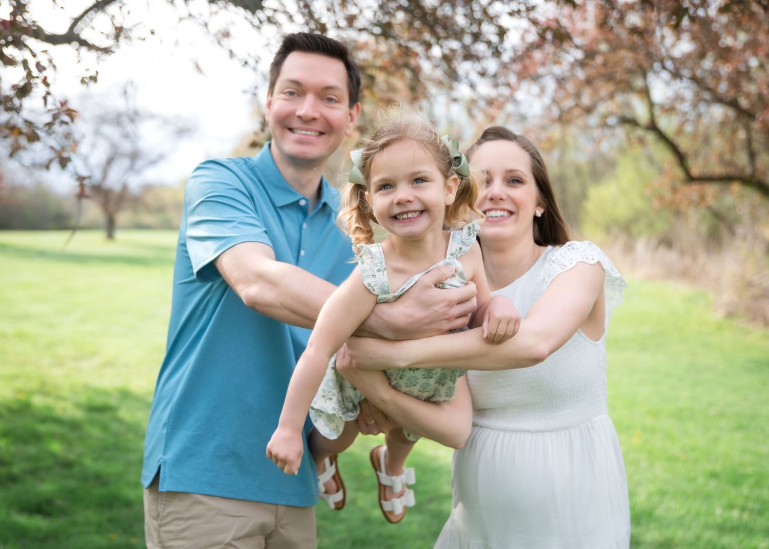 Happy family of three posing outdoors in a lush green park, parents holding their smiling young daughter.
