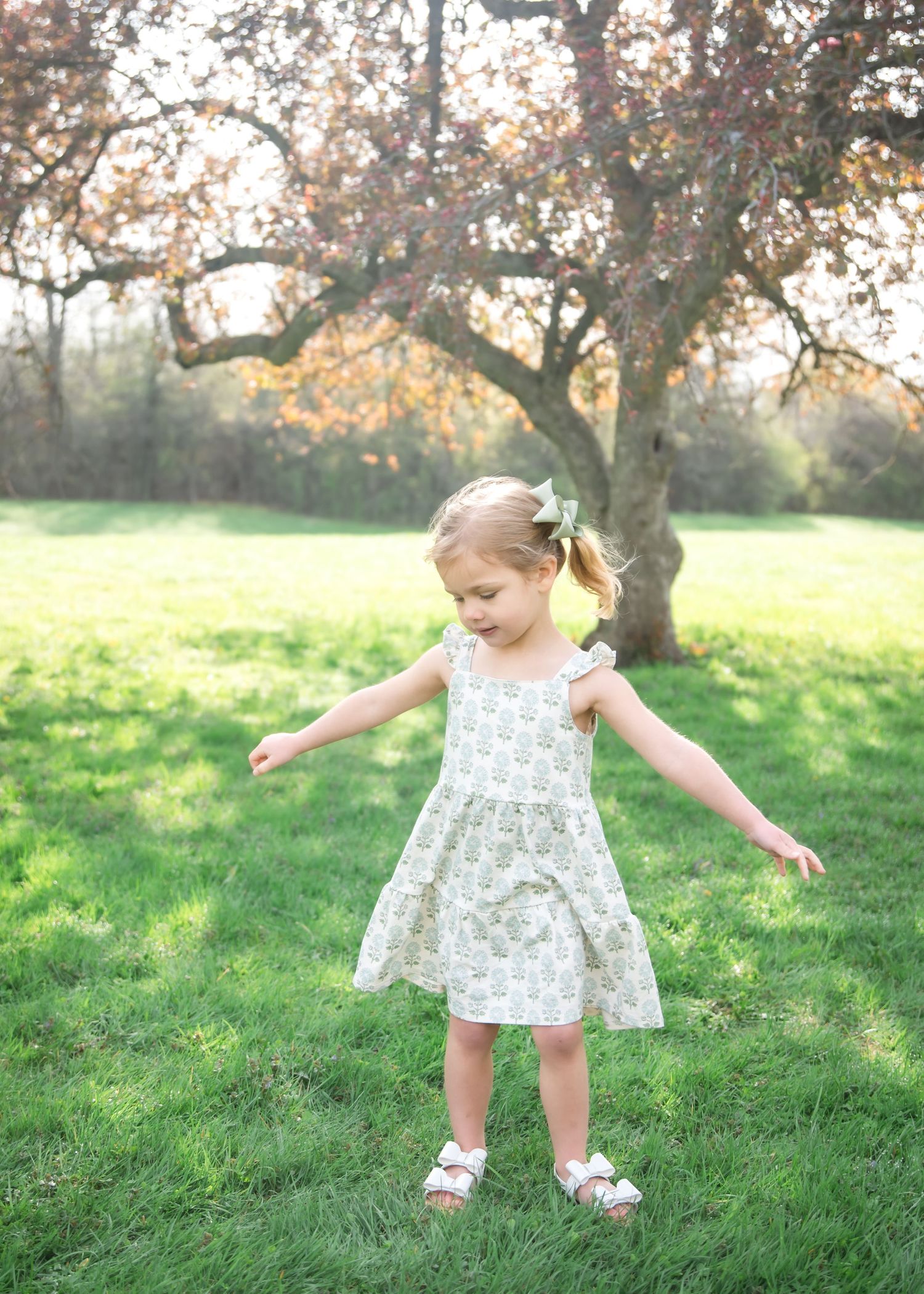 A young girl in a floral dress twirls on green grass with a blooming tree backdrop.