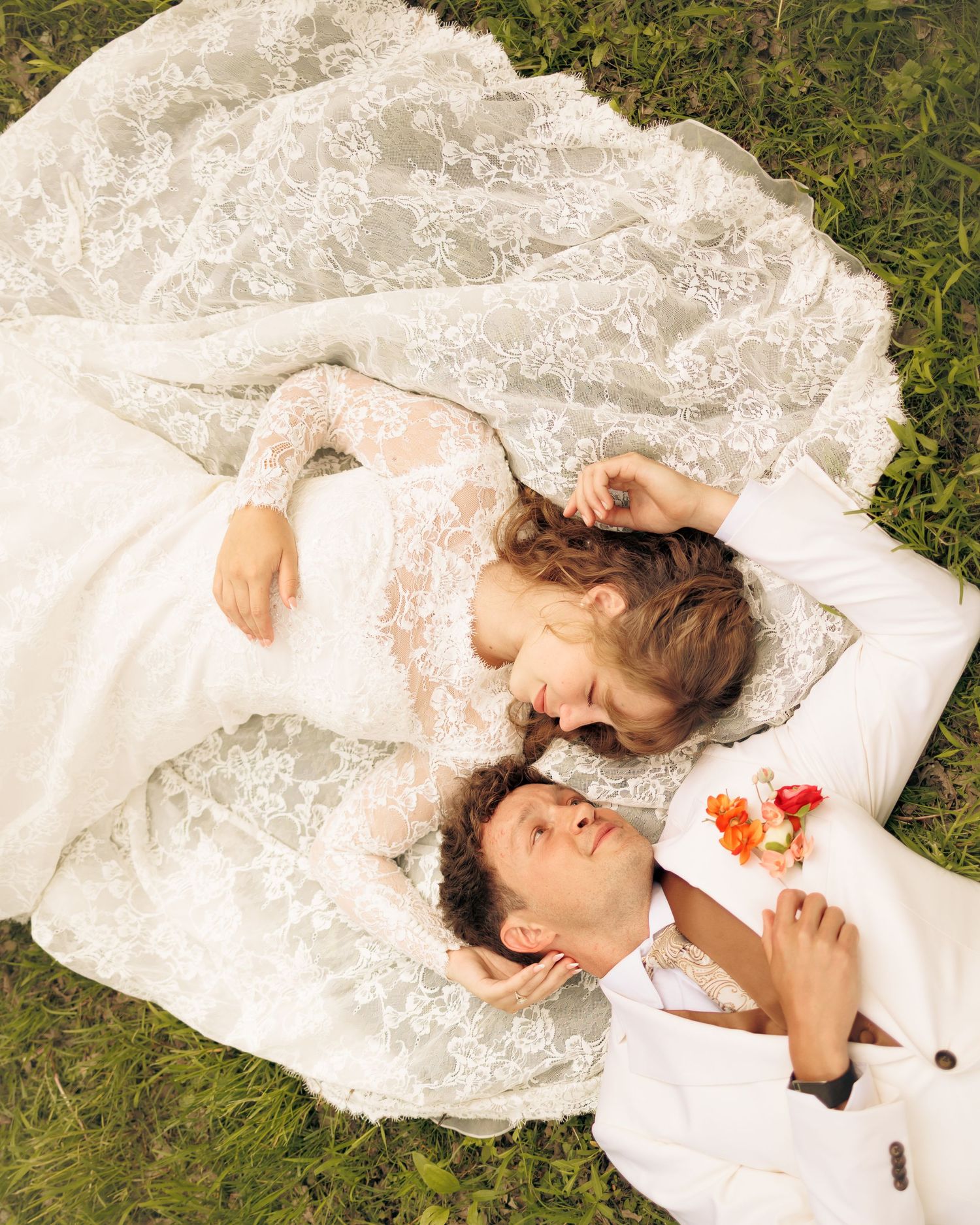 Bride and groom lying on grass, wearing white wedding attire, gazing at each other with colorful flowers.