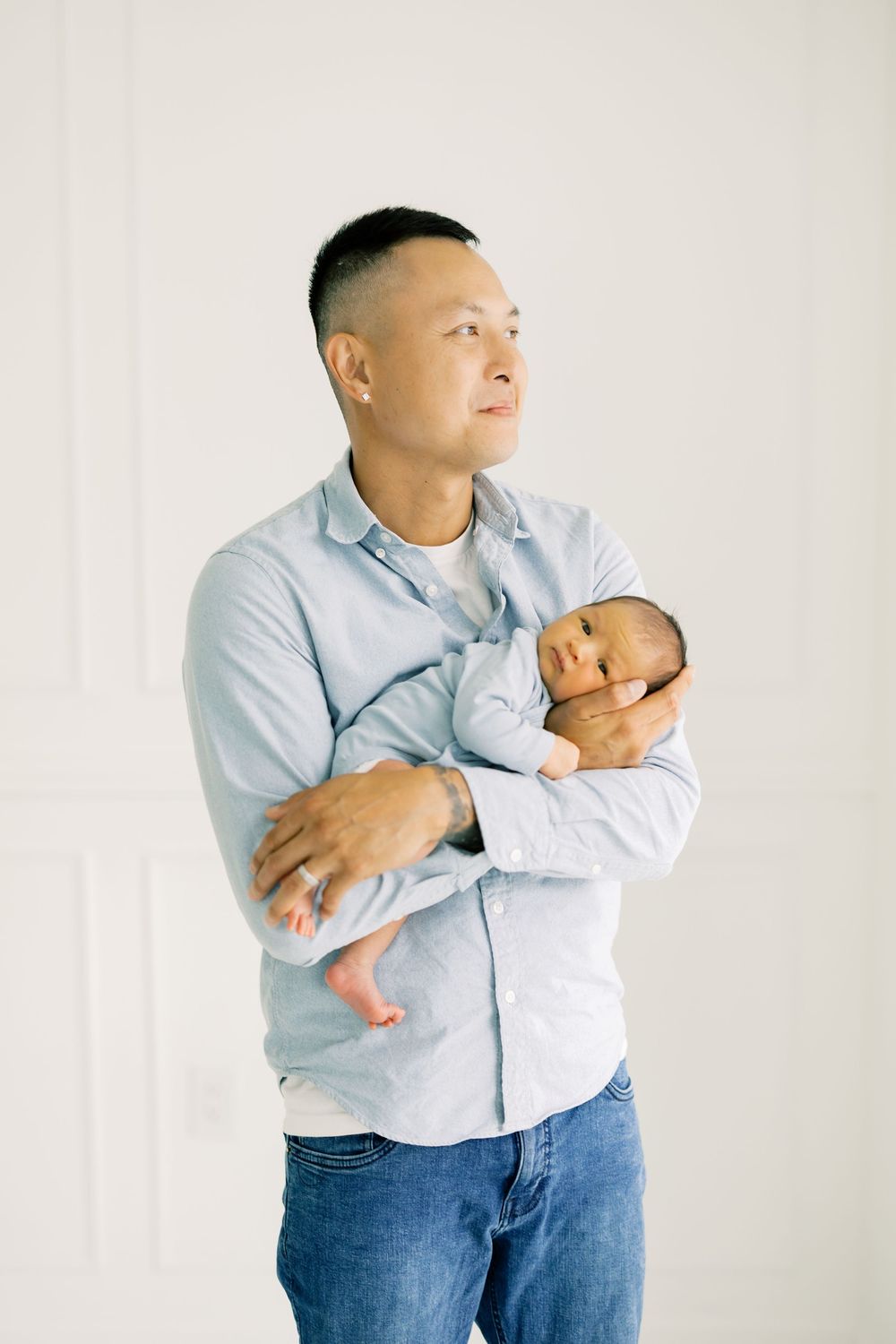 Asian father in blue shirt tenderly holds sleeping newborn baby against his chest in bright white studio.