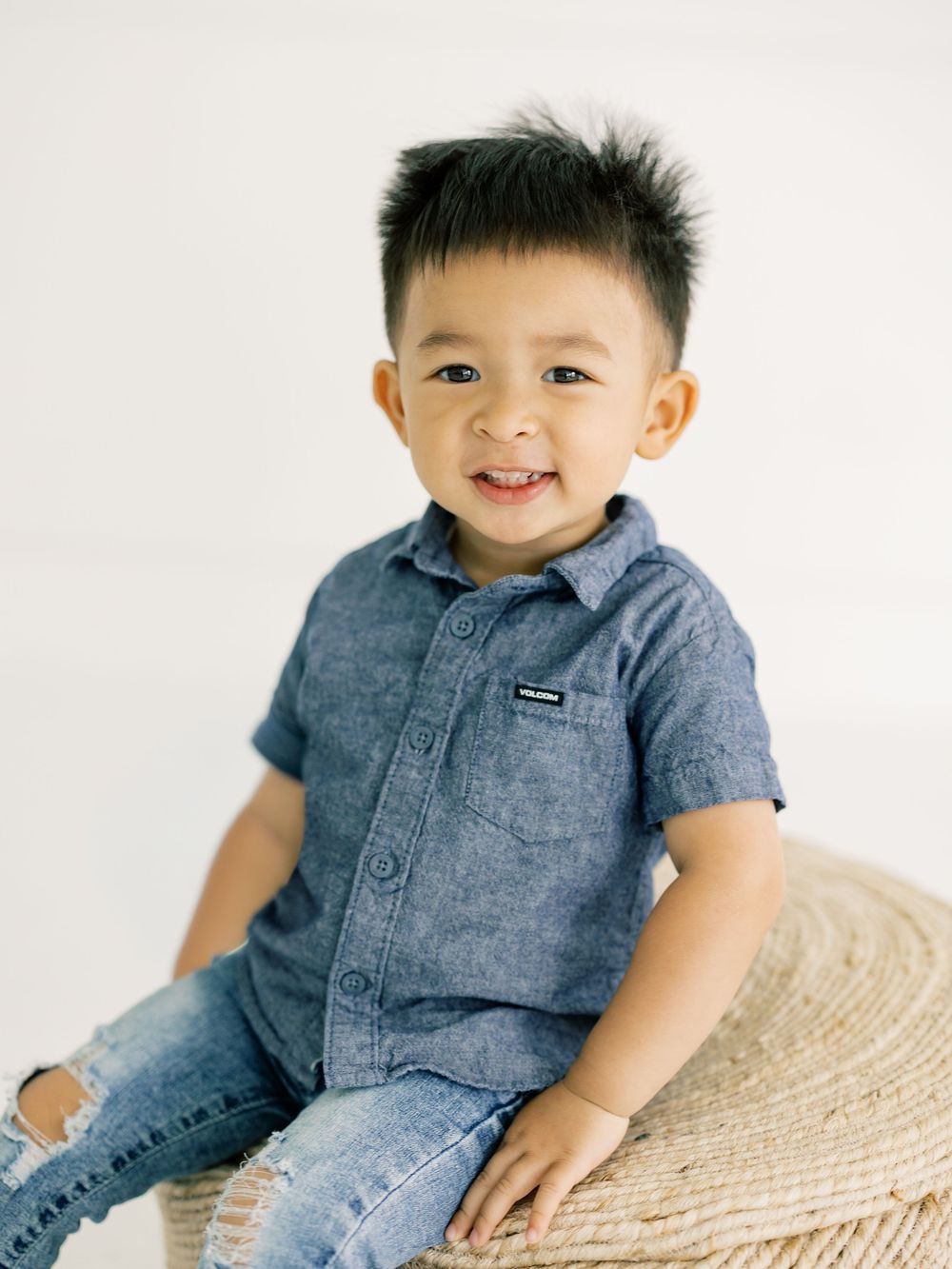 Smiling toddler boy sitting on woven stool wearing blue chambray shirt and jeans in bright white studio.