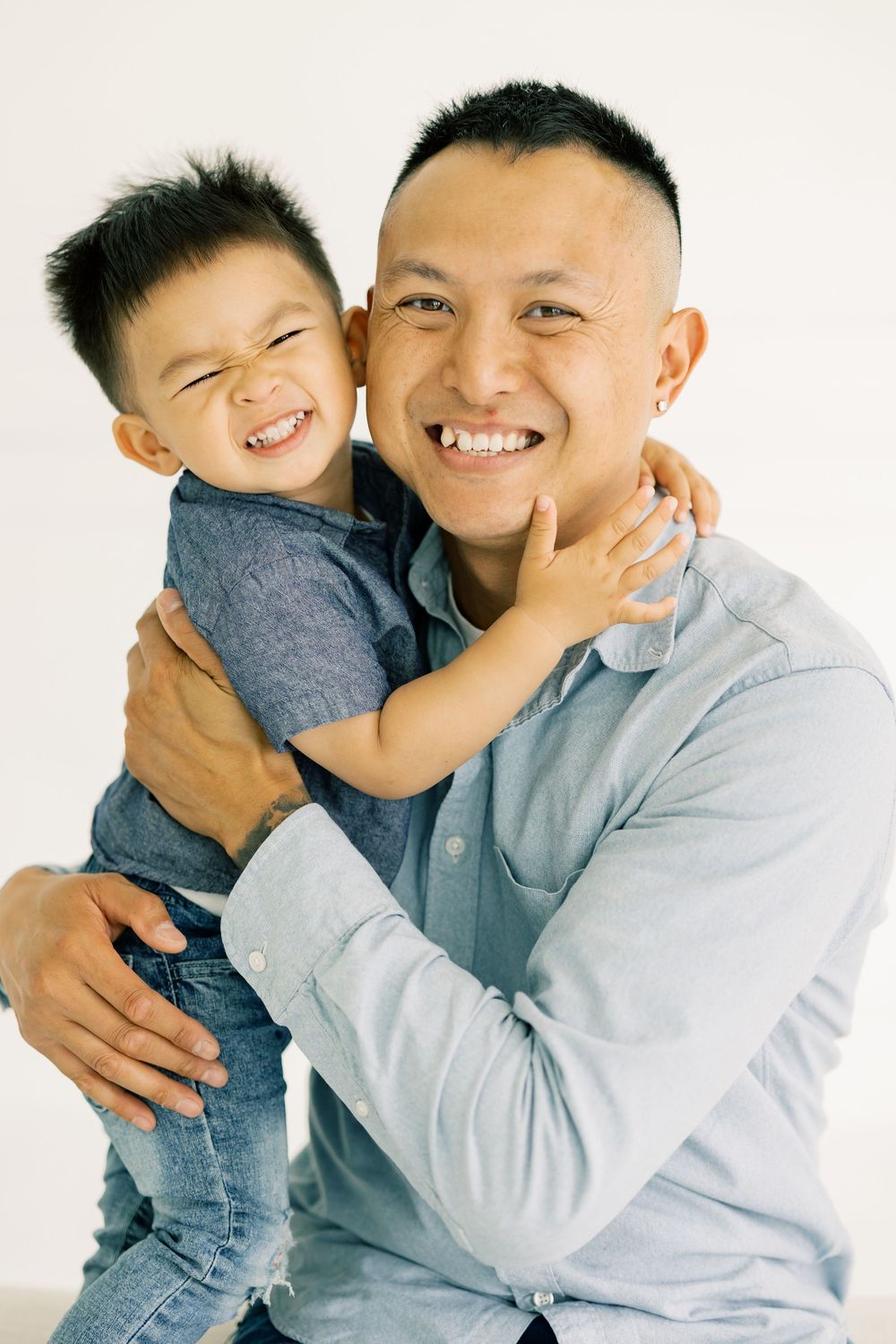 Smiling Asian father holding laughing young son in a warm embrace against a white background.