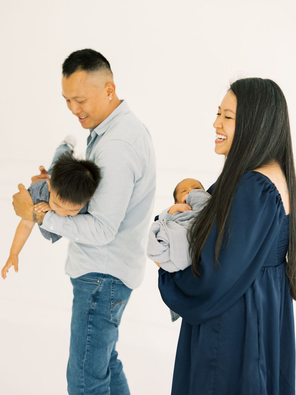 Happy Asian family with father holding toddler and mother cradling newborn in white photography studio.